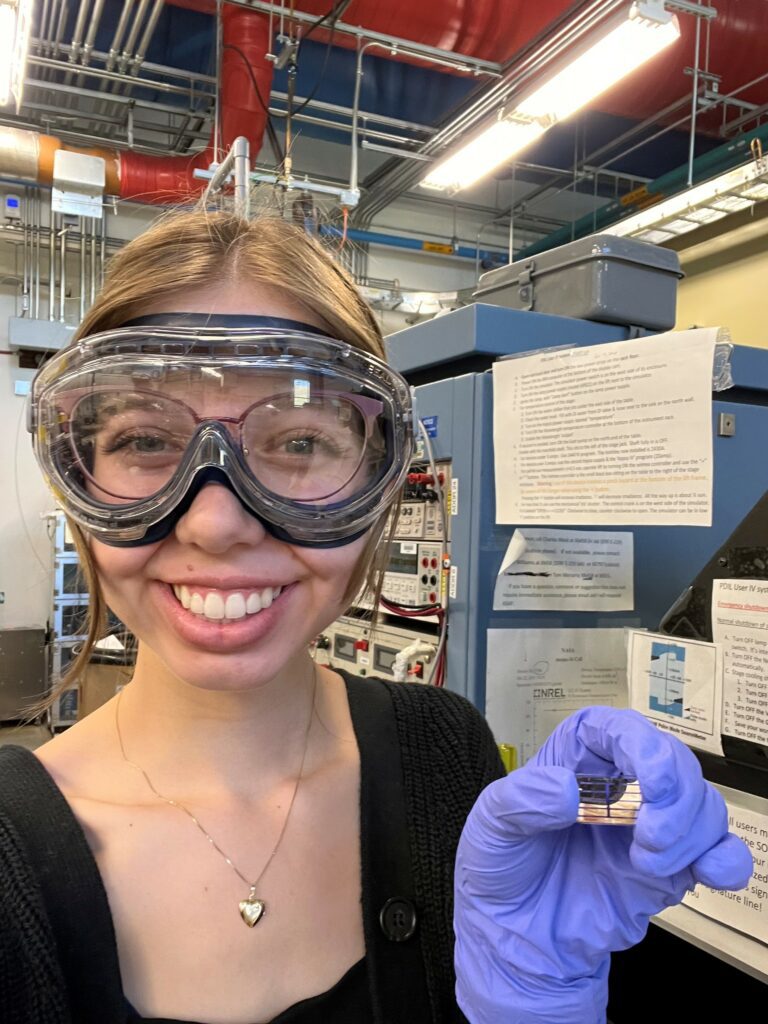 Amanda Behmer in a lab setting, wearing protective goggles and gloves, holding a small device and smiling at the camera.